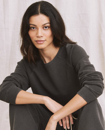 A person with long dark hair and a thoughtful expression sits cross-legged, wearing The Shrunken Sweatshirt in washed black from THE GREAT. CORE KNITS against a neutral backdrop.