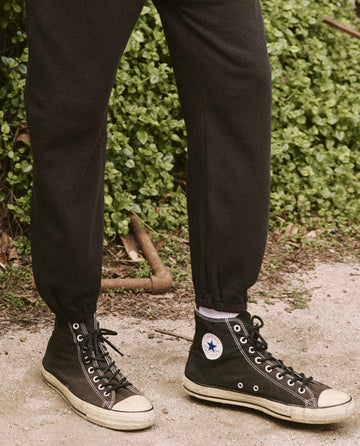 A person wearing black high-top sneakers and The Men's Stadium Sweatpant in Almost Black from THE GREAT. HOL 21 MENS stands on a dirt path with greenery in the background.
