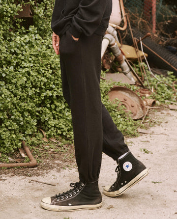 Person wearing The Men's Stadium Sweatpant in Almost Black from THE GREAT. HOL 21 MENS along with black high-top sneakers with white soles, standing on a path next to some greenery and rusty debris.