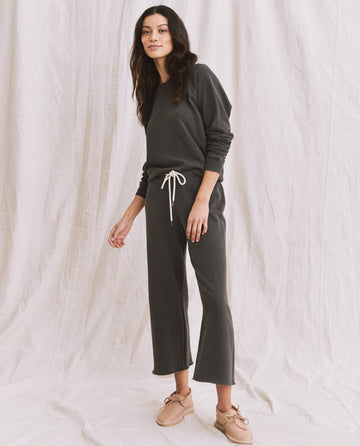 A person stands on a neutral background wearing a dark long-sleeve top, matching pants with a white drawstring, and beige shoes. This ensemble could easily be complemented by The Shrunken Sweatshirt in Washed Black by THE GREAT. CORE KNITS for an added touch of style.