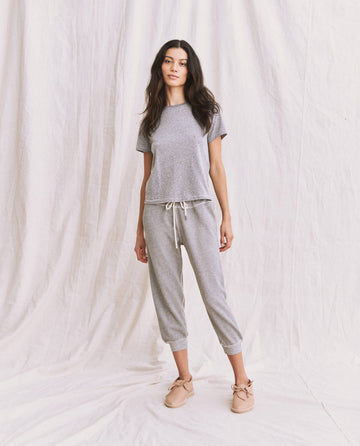 A woman stands in front of a white backdrop, wearing The Little Tee in Heather Grey from THE GREAT. SP22 APRILCAP, paired with matching jogger pants and light-colored shoes.