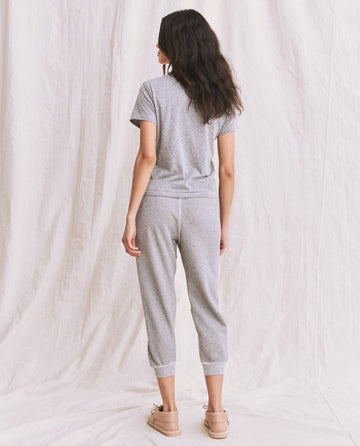 A person with long, dark hair stands with their back to the camera, wearing The Little Tee in Heather Grey and grey jogger pants from THE GREAT. SP22 APRILCAP. The comfortable fit complements the light-colored, crinkled fabric in the background, creating a relaxed vibe.