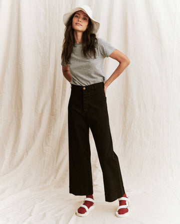 A person stands against a plain backdrop, wearing The Little Tee in Heather Grey from THE GREAT. SP22 APRILCAP, paired with black wide-leg pants, a white bucket hat, and red sandals.