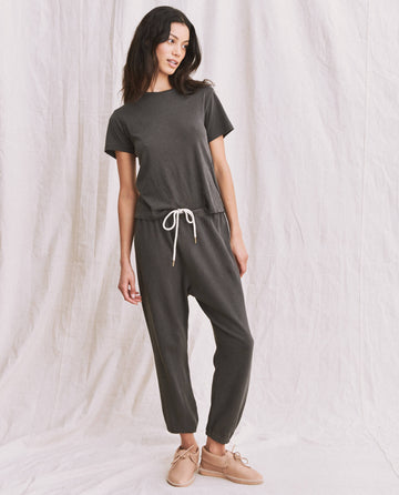 A woman stands against a white background, wearing THE GREAT. SP22 APRILCAP's The Little Tee in Washed Black, made of 100% cotton, and matching drawstring pants. She has long dark hair and holds one hand in her pocket. Her light tan shoes complete the effortlessly stylish look, perfect as a versatile layering piece.