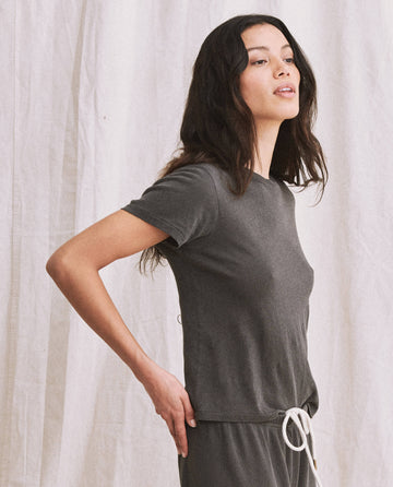 A person stands in a relaxed pose wearing The Little Tee. -- Washed Black, a short-sleeve shirt by THE GREAT. SP22 APRILCAP, paired with matching pants featuring a white drawstring, resembling the simplicity of a vintage men's undershirt, against a white fabric background.