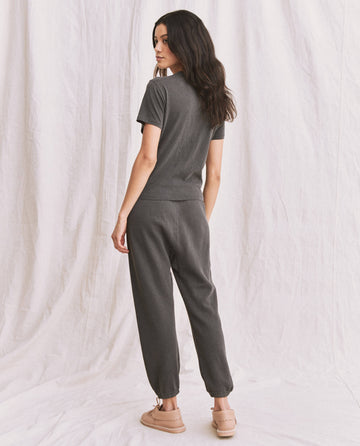 A person with long dark hair, wearing The Little Tee in Washed Black by THE GREAT. SP22 APRILCAP and matching pants, stands turned slightly away against a white fabric backdrop.