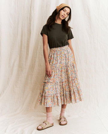 A person stands in front of a white backdrop wearing "The Little Tee" in washed black from THE GREAT. SP22 APRILCAP, paired with a dark green layering piece, a floral tiered skirt, pink socks, beige sandals, and a yellow checkered headband.