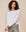 Person with curly hair, wearing The Long Sleeve Crop Tee in TRUE WHITE by THE GREAT. CORE KNITS, paired with gray pants, stands against a neutral background.