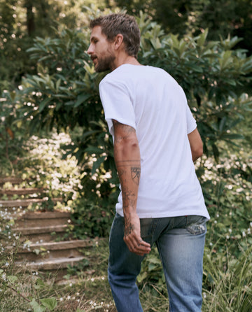 A man in The Men's Slim Tee by THE GREAT. MAN, in True White, and blue jeans walks outdoors on a path surrounded by green foliage.