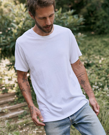 A tattooed man stands outdoors, wearing a plain white "The Men's Slim Tee" in TRUE WHITE by THE GREAT. MAN, paired with light blue jeans. He is looking down with a backdrop of greenery.