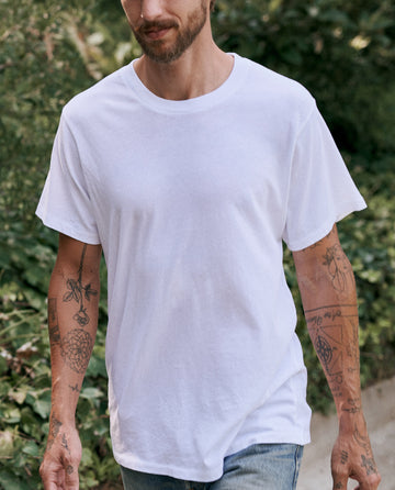 A man is walking outdoors wearing The Men's Slim Tee in True White by THE GREAT. MAN and a pair of jeans. He has tattoos on his arms and is surrounded by greenery.