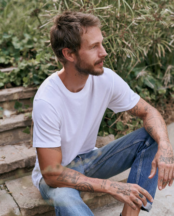 Man with tattoos sits on outdoor steps, wearing The Men's Slim Tee -- TRUE WHITE by THE GREAT. MAN and jeans, surrounded by greenery.