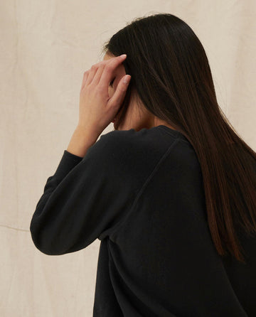 A person with long, straight dark hair is seen from behind, wearing The Shrunken Sweatshirt in Almost Black by THE GREAT. CORE KNITS made from 100% cotton, with one hand raised to their head.