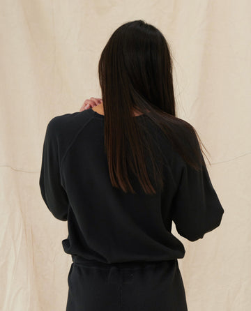 A person with long, straight hair wearing THE GREAT. CORE KNITS' "The Shrunken Sweatshirt" in Almost Black, seen from behind against a light background.