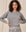 A woman with curly hair wearing The Shrunken Sweatshirt in Varsity Grey from THE GREAT. CORE KNITS stands with her hands on her hips against a cream-colored background.