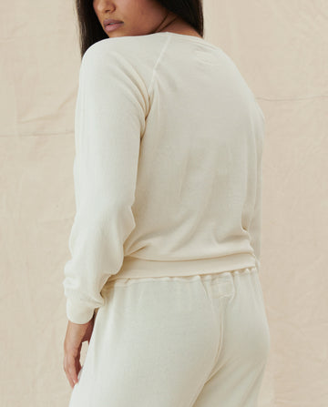 A person is wearing The Shrunken Sweatshirt in washed white and matching pants, standing with their back slightly turned. The outfit evokes the classic simplicity of vintage sweatshirts, effortlessly combining timeless style with modern elegance. This ensemble embodies the distinctive charm and quality of THE GREAT's CORE KNITS collection.