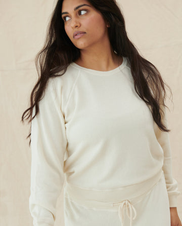 A person with long dark hair, wearing The Shrunken Sweatshirt by THE GREAT. CORE KNITS in washed white and matching 100% cotton pants, looks to the side against a plain beige background.