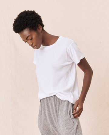 A person wearing The Slim Tee in True White by THE GREAT. CORE KNITS, made from 100% cotton, and gray pants stands against a light background, looking down with one hand in their pocket.