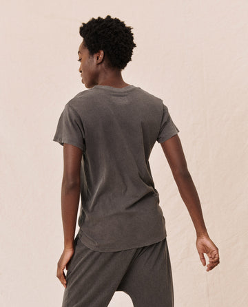 A person with short curly hair is shown from the back, wearing The Slim Tee in Washed Black by THE GREAT. CORE KNITS and matching grey pants. The outfit captures a timeless charm and effortless style.