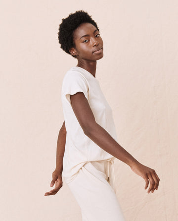 A person with short curly hair is wearing The Slim Tee in Washed White by THE GREAT. CORE KNITS, made from 100% cotton, along with light-colored pants, standing against a neutral background.