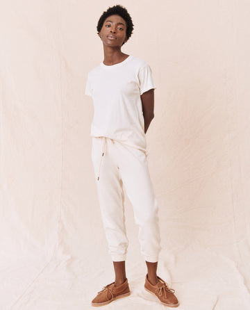 A person stands against a plain background, wearing The Slim Tee in Washed White by THE GREAT. CORE KNITS, white sweatpants, and brown shoes. Their hands are behind their back.