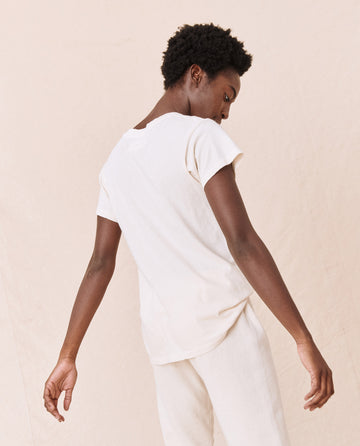 Person standing with back turned, wearing The Slim Tee in Washed White from THE GREAT. CORE KNITS and light-colored pants. The background is plain and neutral.