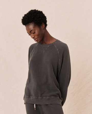 A person with short curly hair stands against a plain background, wearing The Slouch Sweatshirt in washed black from THE GREAT. CORE KNITS and matching pants, looking downward with a gentle smile.