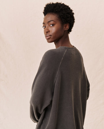 Person with short, curly hair wearing The Slouch Sweatshirt in Washed Black by THE GREAT. CORE KNITS, looking over their shoulder against a plain background.