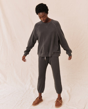 A person is standing against a beige backdrop, wearing The Slouch Sweatshirt in washed black from THE GREAT. CORE KNITS, along with matching pants and brown moccasin-style shoes. The 100% cotton fabric flows naturally as the person with short curly hair looks down, arms slightly out.