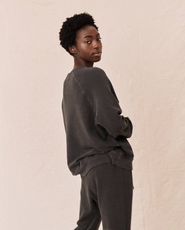 A person wearing The Slouch Sweatshirt in washed black from THE GREAT. CORE KNITS and matching pants stands against a beige background, looking at the camera.