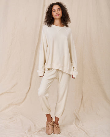 A person stands against a beige backdrop wearing The Slouch Sweatshirt in washed white by THE GREAT. CORE KNITS, made of 100% cotton, along with matching cuffed pants and light tan sneakers.