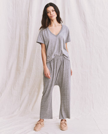 A woman stands against a light-colored backdrop wearing The V-Neck Tee in Heather Grey by THE GREAT. SP22 APRILCAP, styled with matching grey pants and beige shoes.