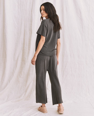A person stands sideways facing away, wearing The V-Neck Tee in Washed Black by THE GREAT. SP22 APRILCAP with wide-leg pants and beige shoes, set against a white fabric backdrop.