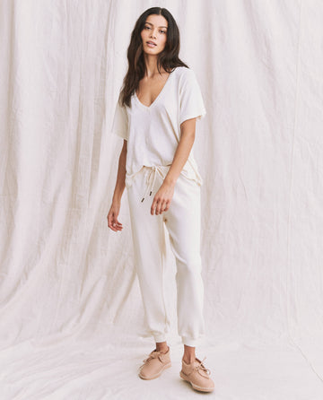 A woman stands against a white fabric backdrop wearing The V-Neck Tee in Washed White from THE GREAT. SP22 APRILCAP, paired with white drawstring pants and beige slip-on shoes.