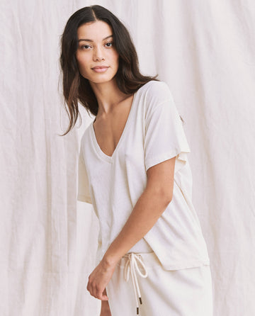A woman with long dark hair is wearing a loose-fitting, short-sleeve, V-neck white top from THE GREAT.'s SP22 APRILCAP collection, specifically "The V-Neck Tee" in Washed White, paired with matching white pants. The boxy fit of her outfit complements the plain white background perfectly.
