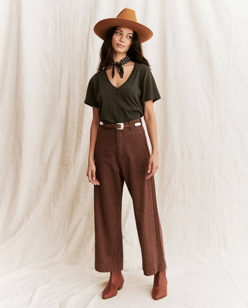 A woman stands against a light backdrop wearing a brown hat, striped brown pants, and brown boots. She is dressed in THE GREAT. SP22 APRILCAP's The V-Neck Tee in Washed Black and has a black bandana tied around her neck.